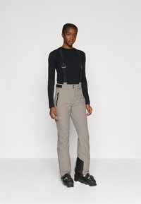 Pantaloni de schi din material gri deschis, cu bretele negre reglabile, trei buzunare și picioare inferioare întărite pentru durabilitate. Botine negre vizibile.