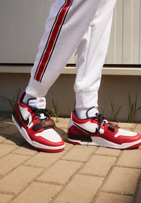 Zapatillas Nike rojas y blancas con acentos negros, lados texturizados y una correa prominente. Llevadas con pantalones blancos que presentan una franja de logo roja.
