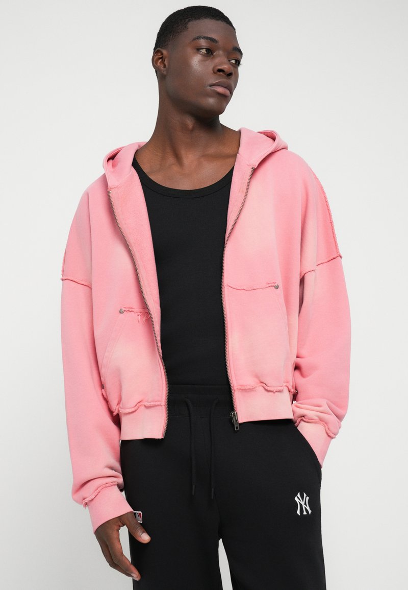 Pink zip-up hoodie with a relaxed fit and raw hem details, worn over a black tank top, paired with black jogger pants featuring logo embroidery.