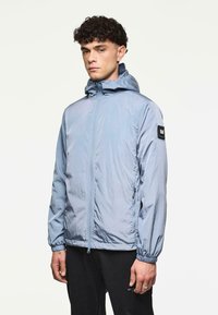 Man met krullend donker haar, gekleed in een lichtblauwe hoodie met rits en zwarte broek, staand tegen een effen lichte achtergrond.