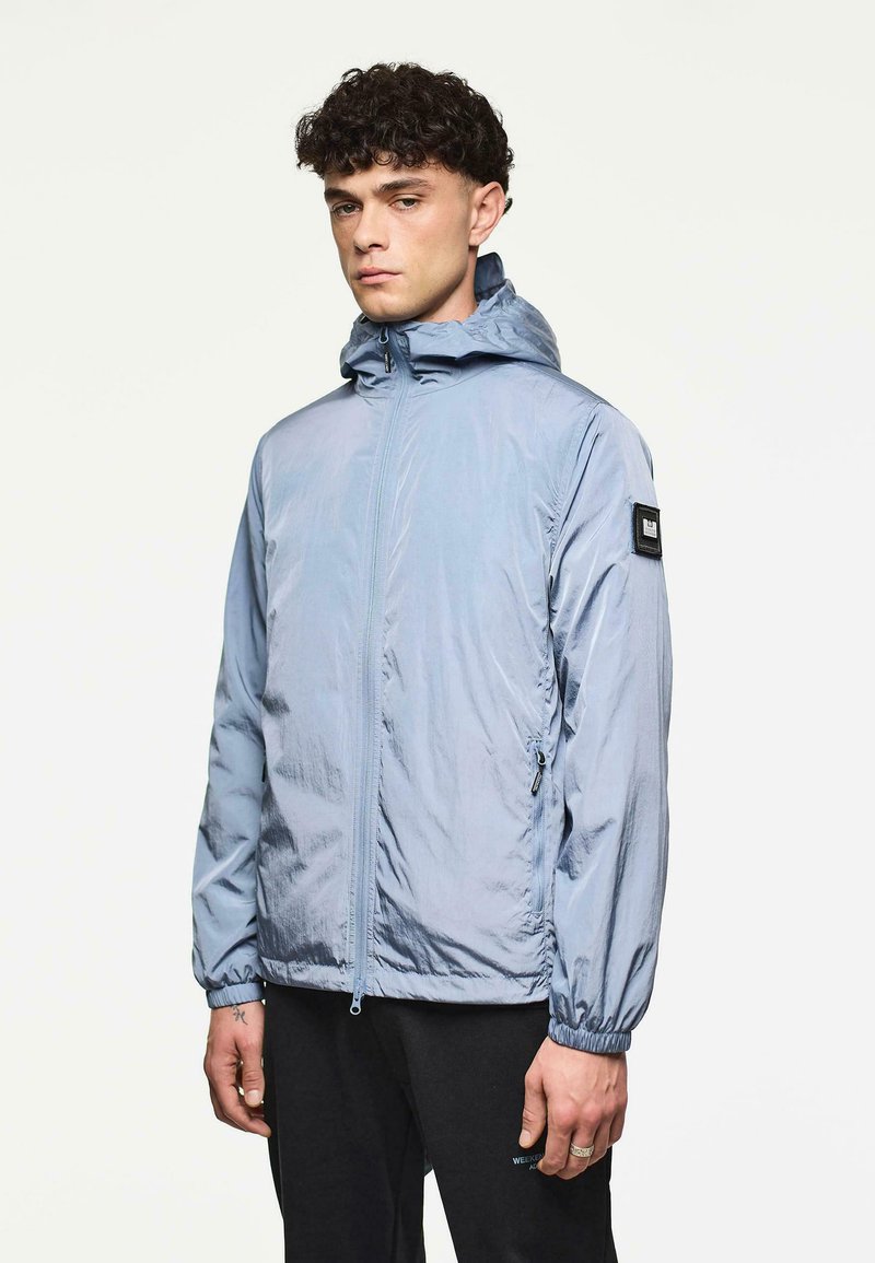 Man met krullend donker haar, gekleed in een lichtblauwe hoodie met rits en zwarte broek, staand tegen een effen lichte achtergrond.