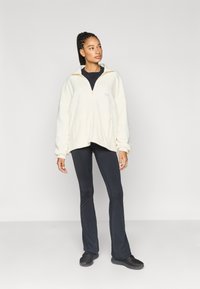 Kermanvärinen fleecejakku vetoketjulla ja korkealla kauluksella, yhdistettynä mustiin liehuviin housuihin ja mustiin kenkiin. Malli seisoo yksivärisen taustan edessä.