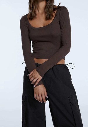 Mujer lleva un top corto marrón oscuro ajustado de manga larga y pantalones cargo negros sueltos con cintura con cordón, manos cruzadas delante.