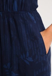 Navy blue fabric with subtle dark leaf and geometric patterns, elastic waistband, and hand resting inside side pocket.