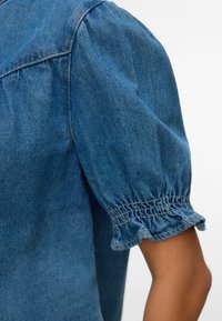 Manche de chemise en denim avec un poignet élastique froncé et un détail subtil d’épaule bouffante sur un bras à la peau hâlée.
