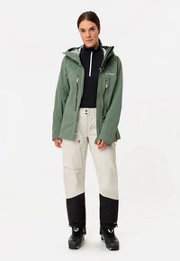 Grüne wasserdichte Jacke mit zwei Reißverschlusstaschen, einer Kapuze und schwarzen Akzenten, kombiniert mit hellen Hosen und schwarzen Skischuhen.