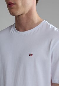 White cotton t-shirt featuring a small red and blue Norway flag embroidery on the left chest. The design is simple with a crew neck.