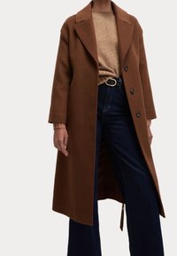 Brązowy wełniany płaszcz z zauważonym kołnierzem, trzema guzikami i bocznymi rozcięciami. Nosi się go na beżowym swetrze i ciemnoniebieskich dżinsach z paskiem.
