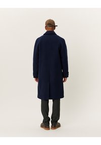 Ναυτικό κάπρο από μαλλί με ίσια γραμμή, διπλές κουμπότρυπες και προσαρμοσμένο γιακά. Φορεμένο πάνω από γκρίζες παντελόνες και καφέ παπούτσια.