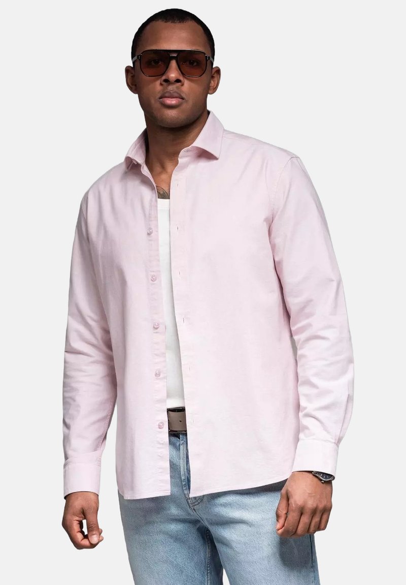 Lichtroze overhemd met knopen van katoen, gecombineerd met een witte tanktop, lichtblauwe jeans en oversized bruine zonnebril.