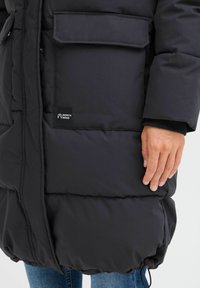 Svart pufferjakke med glidelås, to frontlommer, elastiske mansjetter og et logopatch på den nedre siden, laget av quiltet stoff.