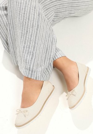 Piedi che indossano ballerine intrecciate beige chiaro con piccoli fiocchi, abbinate a pantaloni larghi a righe grigie e bianche.