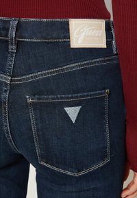 Temno modri traper hlače z velikim Guess logotipom, ki imajo belo šivanje in svetlo modri trikotni poudarek na zadnjem žepu.