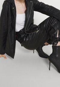 Veste en simili cuir noir avec fermeture éclair à l'avant, associée à un pantalon plissé noir et des bottines. La tenue présente des textures élégantes et des coupes asymétriques.