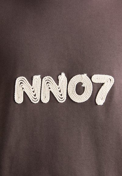 Tissu marron foncé avec broderie "NN07" en fil de couleur crème torsadé. Les lettres ont une apparence texturée, semblable à de la corde.