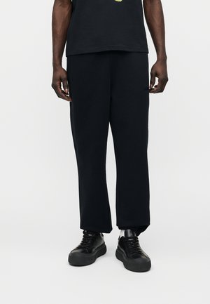 Mężczyzna w czarnych spodniach o prostych nogawkach i czarnych sneakersach, stojący na tle jednolitego białego tła.