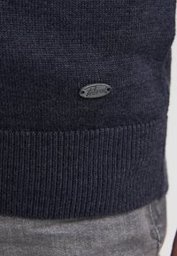 Pulover tricotat de culoare bleumarin, cu un tiv cu nervuri. O etichetă cu logo este coaptă pe față. Textura este netedă, cu cusături uniforme.