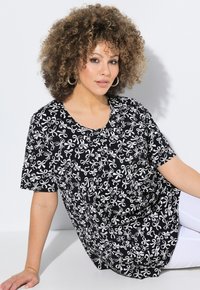 Schwarzes Shirt mit weißen Blumen- und Schleifenmustern, kurze Ärmel, runder Ausschnitt, lockere Passform, kombiniert mit weißen Hosen.
