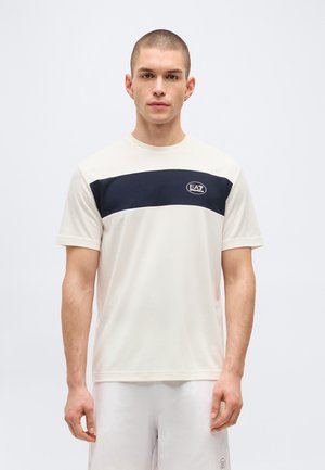 Fiatal férfi rövid hajjal, fehér pólót visel fekete vízszintes csíkkal és EA7 logóval, valamint fehér rövidnadrágot, egy egyszínű háttér előtt áll.