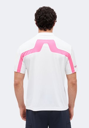 Hombre con cabello rizado oscuro que lleva una camisa blanca de manga corta con franjas geométricas rosas en la espalda y las mangas, de espaldas.