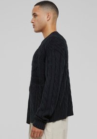 Pull en maille torsadée noir avec un motif texturé, des poignets côtelés et une coupe décontractée, porté par une personne de profil.