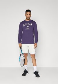 Lilla pikkseeliku varrukatega Adidas särk valge logoga, millele on sobitatud valged lühikesed püksid lilla äärega. Mustad tennised ja tennisereket.