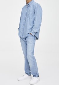 Hellblaues Jeanshemd mit Knopfleiste am Kragen und Tasche, kombiniert mit hellblauen Jeans und weißen Turnschuhen. Glatte Textur, lässige Passform.