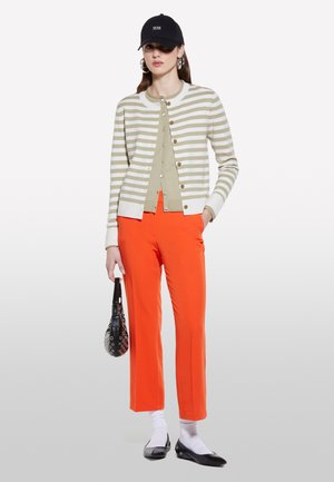 Cardigan a righe beige chiaro e crema, abbinato a pantaloni arancioni brillanti. Gli accessori includono un berretto nero e una piccola borsa strutturata.