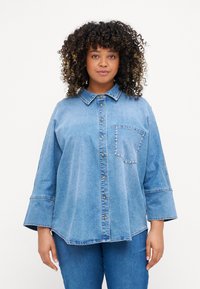 Camisa de ganga azul clara com gola, bot�ões frontais e um bolso no peito decorado com tachas prateadas. Apresenta mangas largas e um corte descontraído.