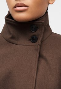 Κοντινό πλάνο ενός ατόμου που φοράει ένα καφέ παλτό με ψηλό γιακά, κλειστό με δύο μεγάλα μαύρα κουμπιά, εμφανίζοντας τα χείλη και μέρος του προσώπου.