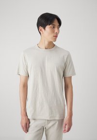 T-shirt beige clair à col rond, fabriqué en tissu doux et texturé. Doté de manches courtes et d'un petit logo sur la poitrine. Design simple.
