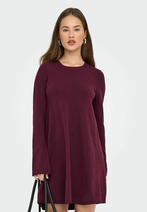 Femme portant une robe longue à manches, de couleur violet foncé, tenant les poignées d'un sac à main noir devant un fond clair.