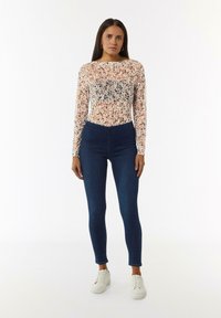 Kwiatowy bodysuit z długim rękawem w różowe, zielone i niebieskie wzory; ciemnoniebieskie jeansy z wysokim stanem typu skinny; połączone z białymi sneakersami.