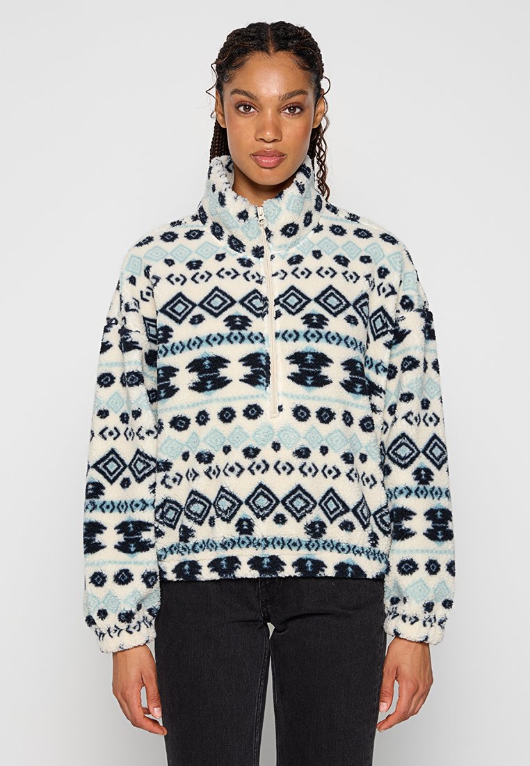 Pull en polaire crème avec un motif géométrique bleu et marine. Doté d'un col montant et d'une fermeture éclair sur le devant. Tissu doux et texturé.