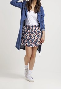 Woman wearing a white top, floral skirt, denim jacket, white socks, and white sneakers, standing against a plain background.