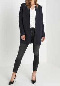 Marinefarbener strukturierter langer Cardigan mit offener Vorderseite, kombiniert mit einer weißen Bluse und schwarzen Skinny Jeans, abgerundet mit spitzen schwarzen Absätzen.
