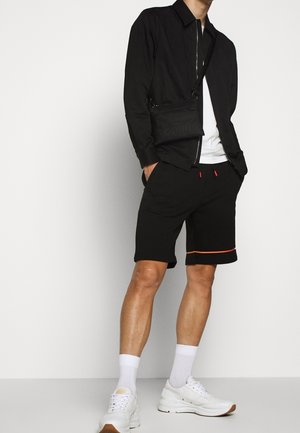 Homme portant une veste noire zippée, un t-shirt blanc, un short noir avec une bordure orange, des chaussettes blanches, des baskets blanches et un sac bandoulière noir.