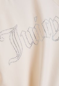 Top beige chiaro con un tessuto testurizzato, decorato con borchie argentate fissate a caldo che formano la parola "Juicy" in una scrittura decorativa.