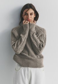 Lichtbruine gebreide trui met een gestructureerd patroon, geribbelde manchetten en onderste zoom. Model houdt de kraag vast, staand tegen een effen achtergrond.