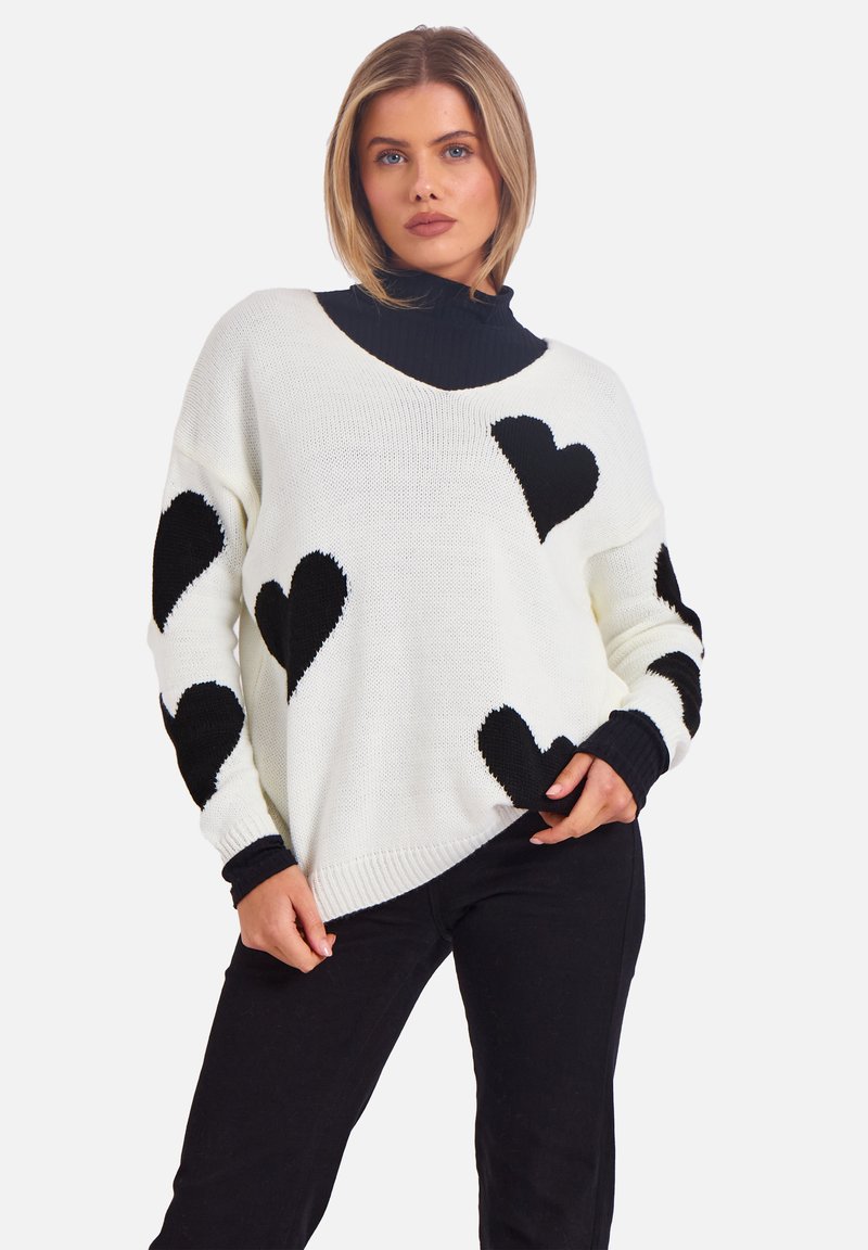 Weißer Strickpullover mit schwarzen Herzmustern, V-Ausschnitt und gerippten Bündchen, getragen über einem schwarzen Rollkragenpullover und kombiniert mit schwarzen Hosen.