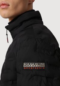 Chaqueta acolchada negra con cuello alto. Cuenta con un logo bordado de "Napapijri geographic" en el hombro. El material parece suave y texturizado.