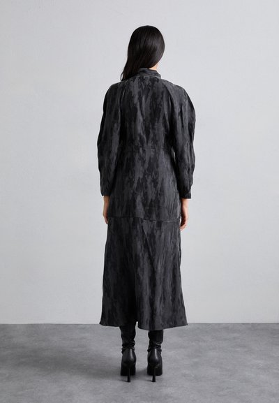 Zwarte, mid-length jurk met een getextureerd patroon en pofmouwen, met een hoge hals en een vloeiende rok. Draagt met aansluitende zwarte laarzen.