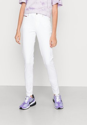 Persoon draagt witte slim-fit jeans, lichtpaars tie-dye shirt en paarse sneakers met witte veters, staand tegen een effen grijze achtergrond.