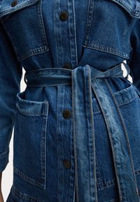 Giubbotto in denim con lavaggio blu, dotato di ampie tasche frontali, chiusura a bottone e una cintura in tessuto abbinato legata in vita.