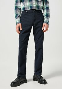Mörkblå byxor med slim fit, tillverkade av vävt tyg. Har framfickor och en ren midja, ihopparade med svarta skor.