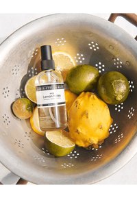 Bottiglia spray in vetro trasparente etichettata "Lemon Notes" accanto a limoni a fette, lime e un frutto giallo in uno scolapasta di metallo forato.