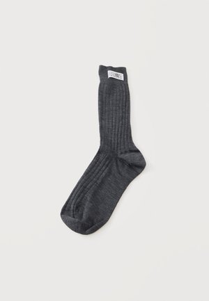 Calcetín vestido de color gris oscuro con costillas, con una etiqueta rectangular blanca cerca del puño, colocado plano sobre un fondo blanco liso.