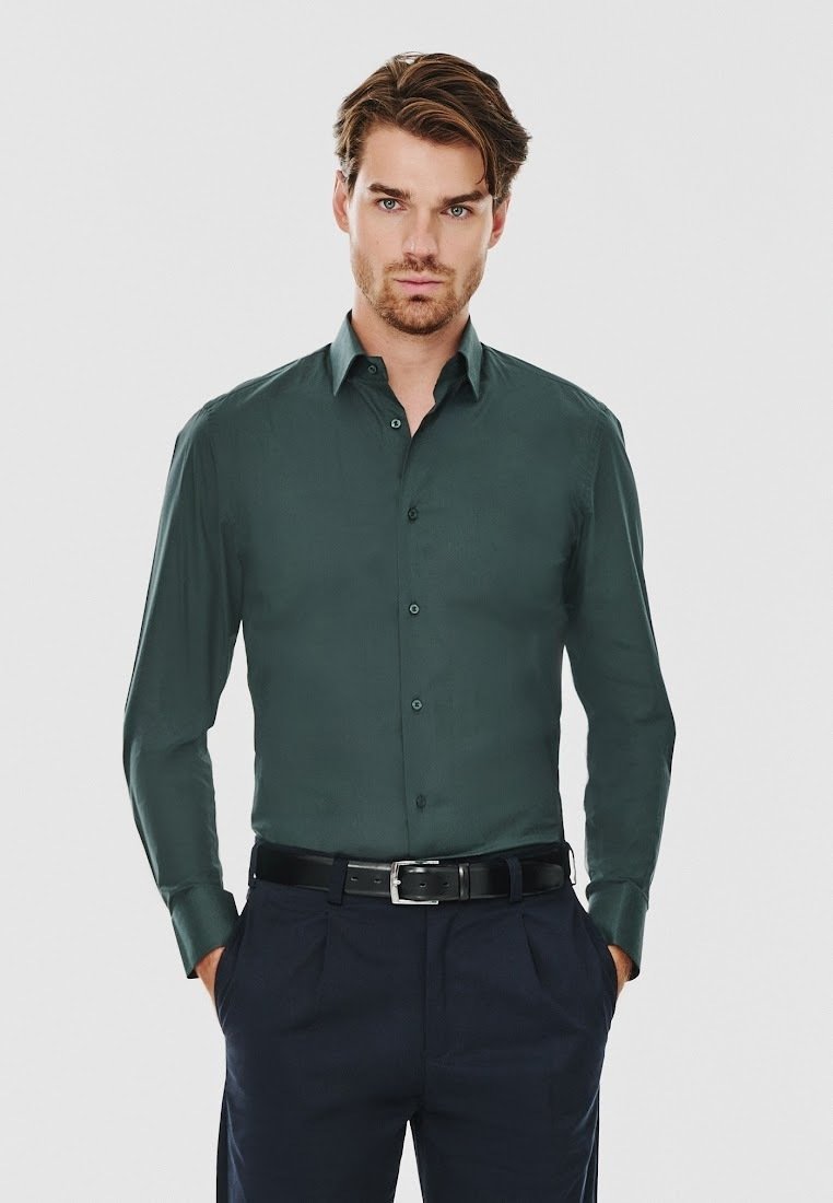 Ciemnozielona, długa koszula z guzikami, wykonana z gładkiego materiału, z pointed collar i dopasowanym krojem, zestawiona z ciemnymi spodniami i paskiem.