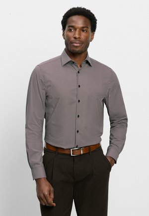 Uomo che indossa una camicia a maniche lunghe con fantasia e bottoni, pantaloni scuri e cintura marrone, in piedi con una mano in tasca davanti a uno sfondo semplice.