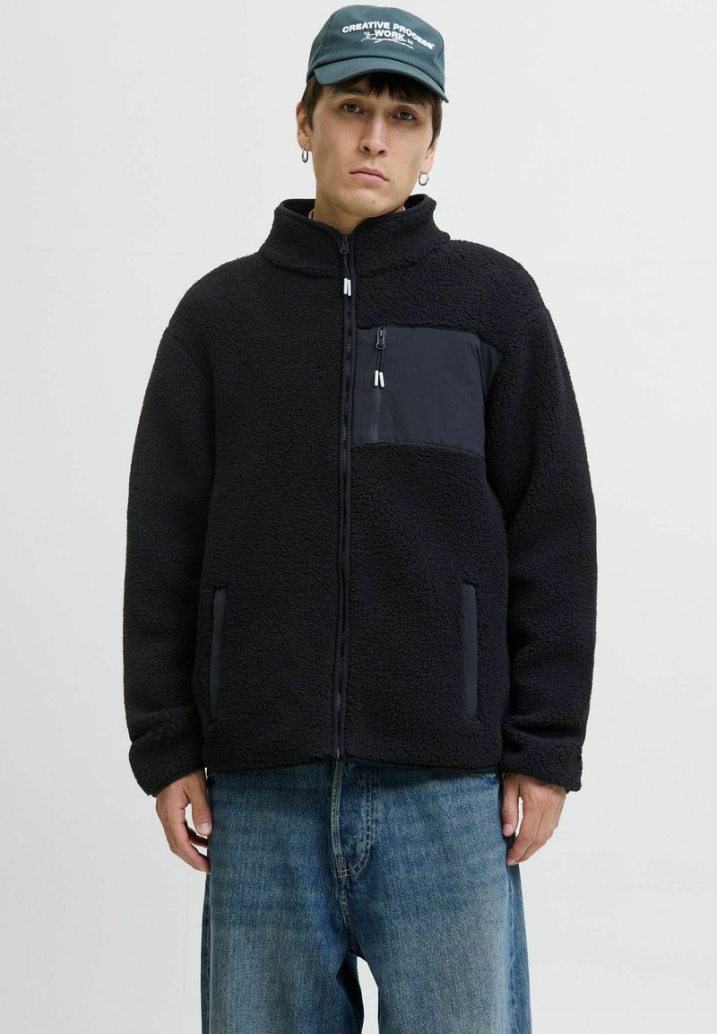 Fleecejacke in Schwarz mit strukturiertem Stoff, Reißverschlusstasche auf der Brust, seitlichen Reißverschlusstaschen und Stehkragen. Trägt eine Kombination mit blauen Jeans.